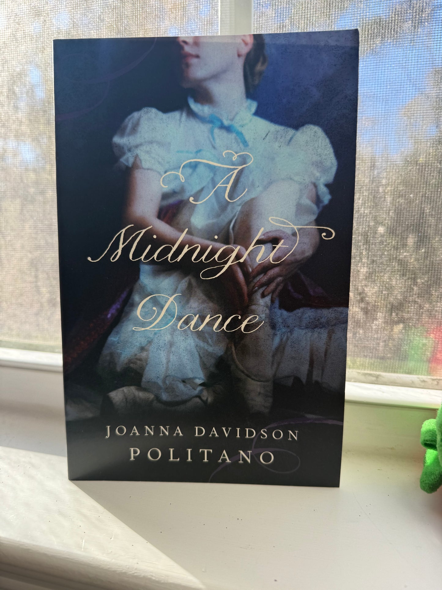 A book cover of 'A Midnight Dance' by Joanna Davidson Politano, featuring a woman in a white and blue dress on the cover, set against a backdrop of a window with a clear sky.