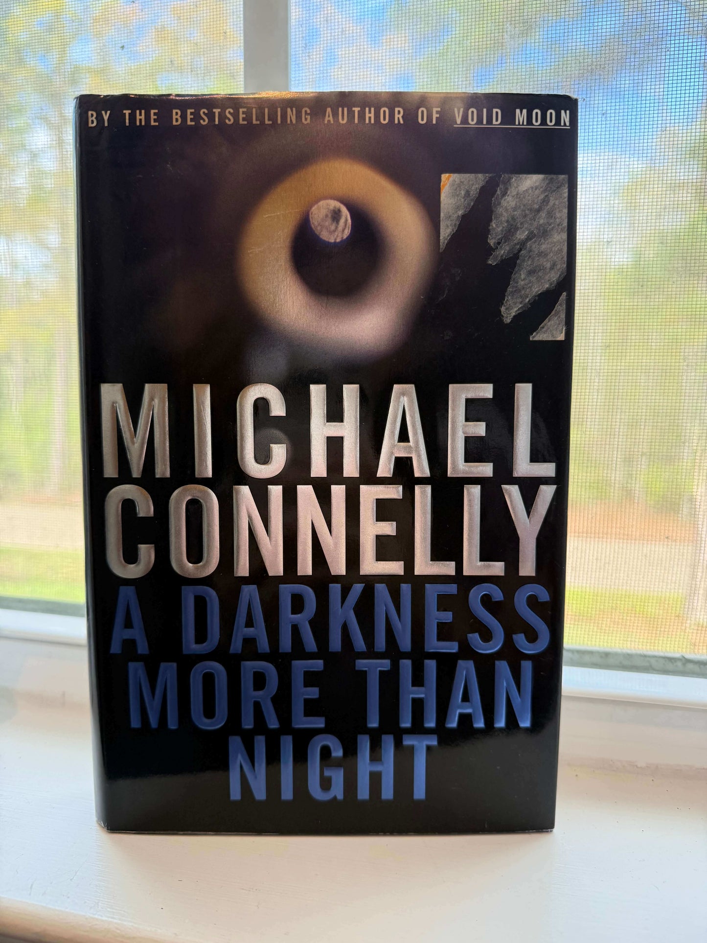 Book titled 'A Darkness More Than Night' by Michael Connelly on a windowsill with a blurred outdoor background.