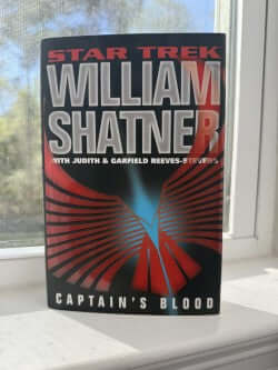 Book titled 'Captain's Blood' by William Shatner on a windowsill with a blurred outdoor background