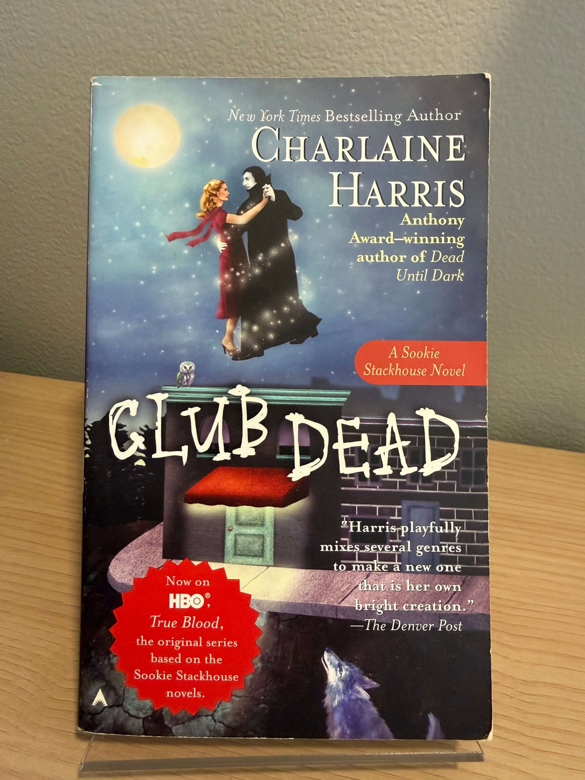 A paperback copy of the book 'Club Dead' by Charlaine Harris, featuring a dark, spooky cover illustration with a couple floating in a starry night sky, a red house, and a red circular promotional badge.
