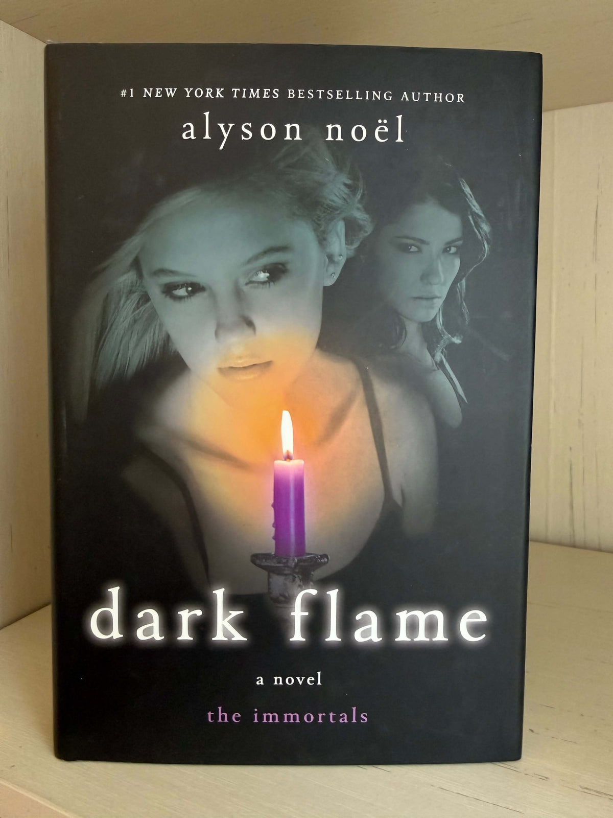 The book cover of 'Dark Flame: A Novel' by Alyson Noel, featuring a dark background with a glowing purple candle and two women's faces, photographed standing upright on a light wood surface.