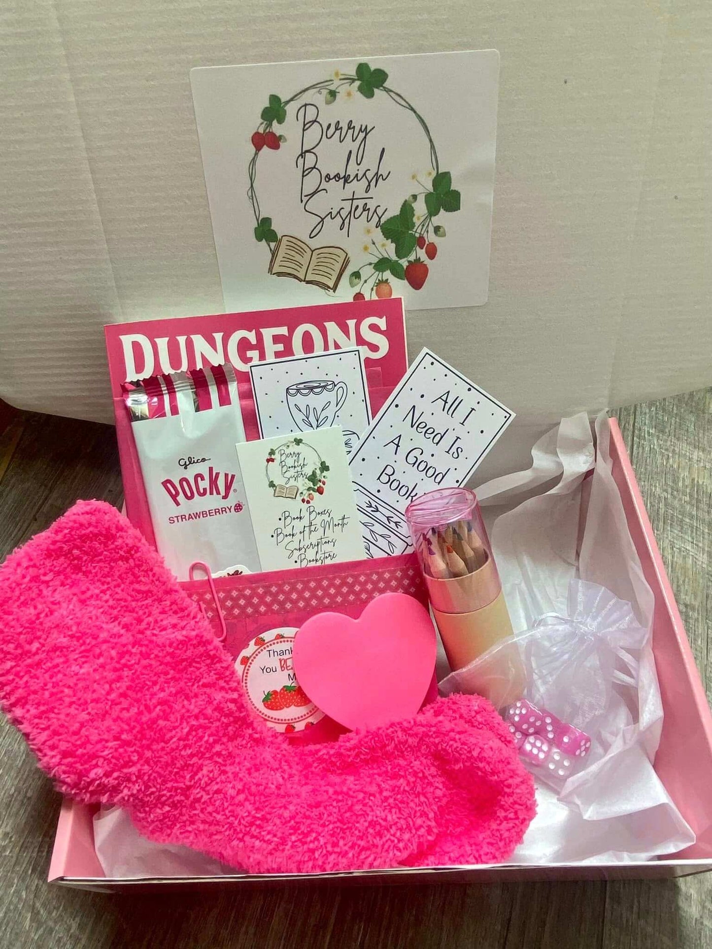 Pink gift box with a book, snacks, and other items on a wooden surface.
