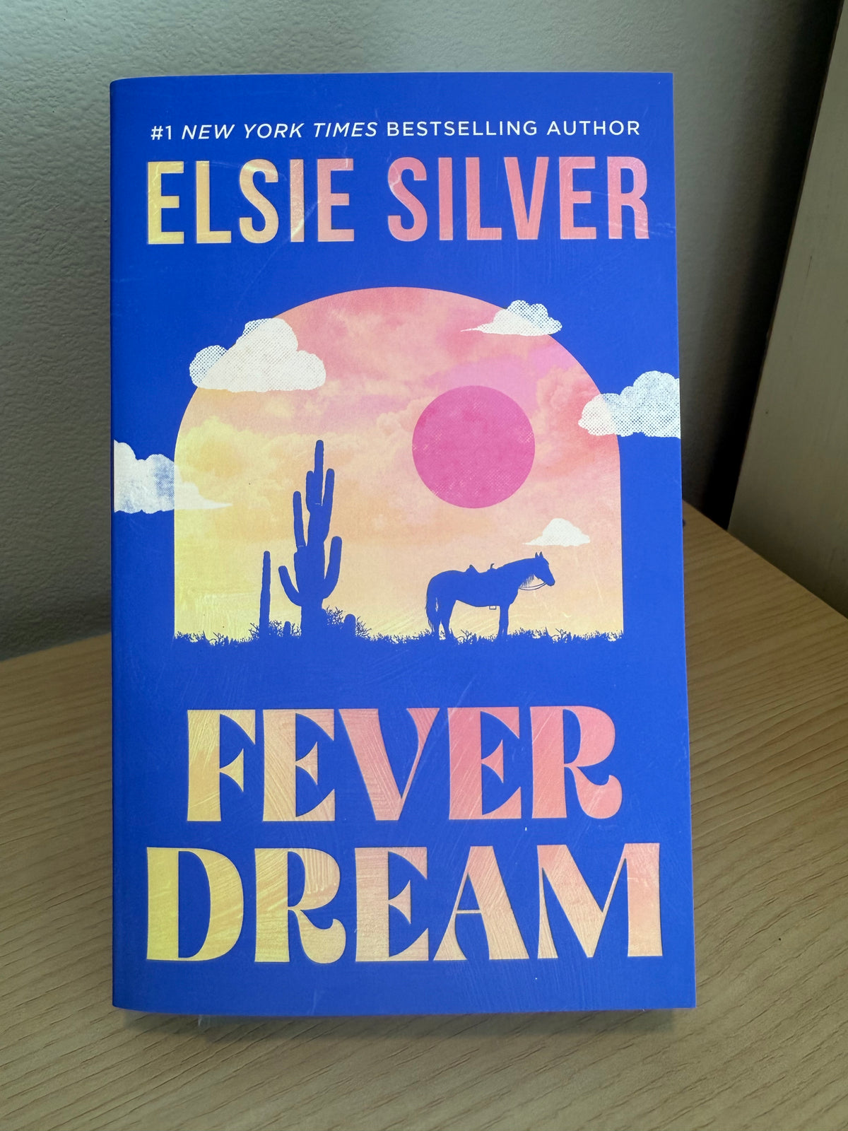 Book cover of 'Fever Dream' by Elsie Silver, featuring a blue background with a desert sunset illustration and stylized title text, photographed standing on a wooden surface against a neutral wall.