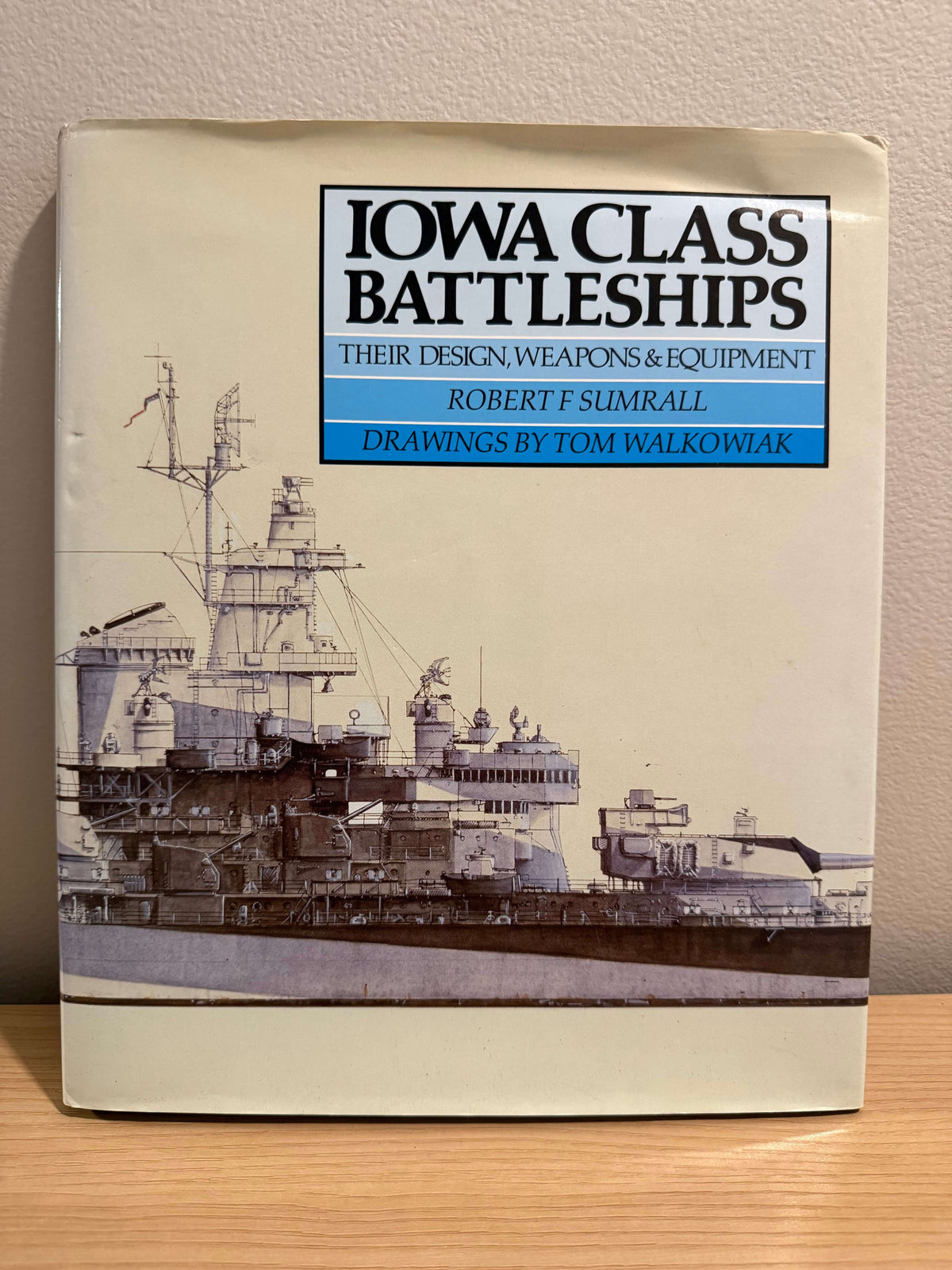 A book titled 'Iowa Class Battleships' by Robert F. Sumrall, with drawings by Tom Walkowiak, standing on a wooden surface against a beige wall.