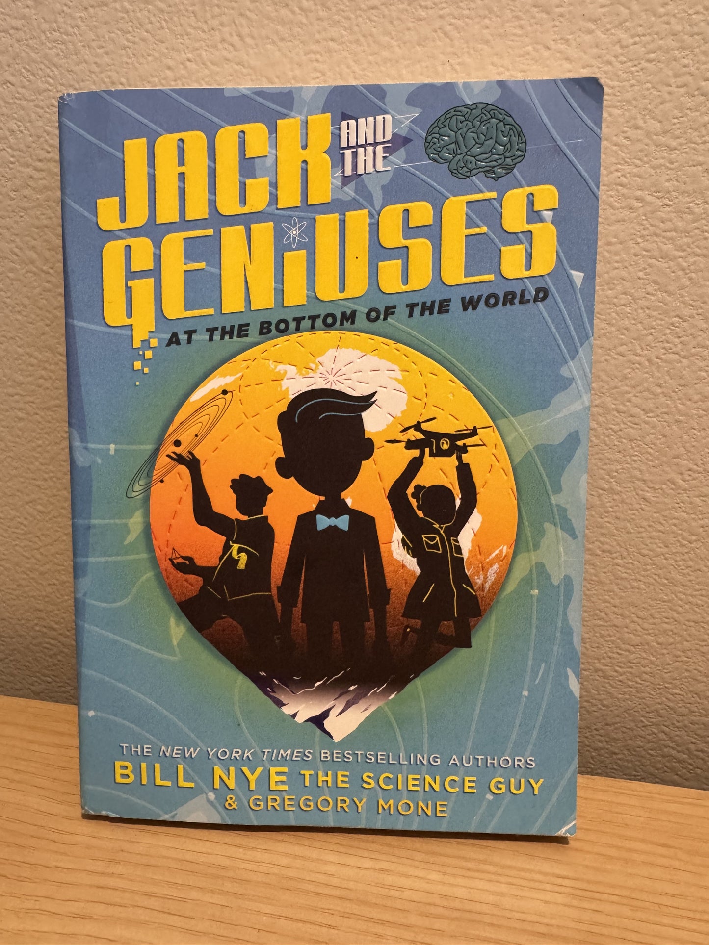 The book 'Jack and the Geniuses: At the Bottom of the World' by Bill Nye and Gregory Mone, standing upright on a wooden surface against a beige wall.