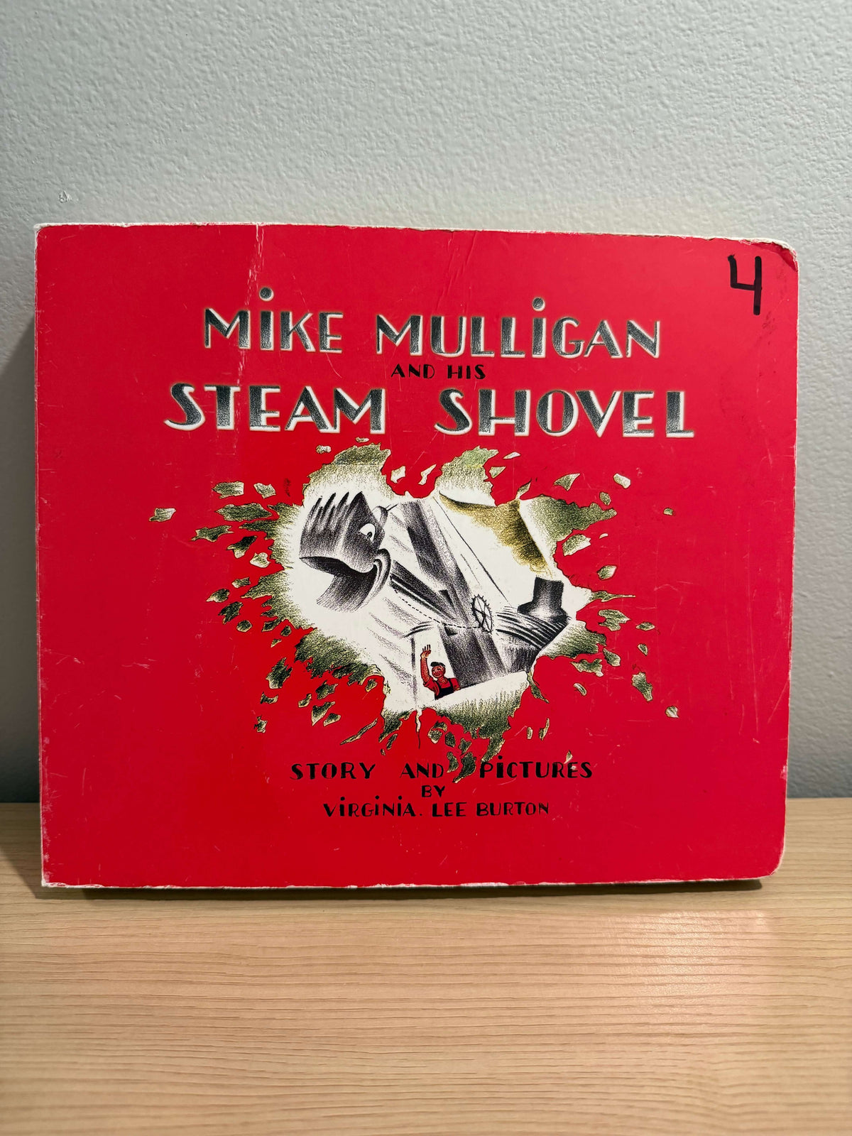 Vintage children's book titled "Mike Mulligan and His Steam Shovel" by Virginia Lee Burton, featuring a bright red cover with a central illustration of a steam shovel and the title in large white letters, photographed standing on a light wood surface against a gray wall.