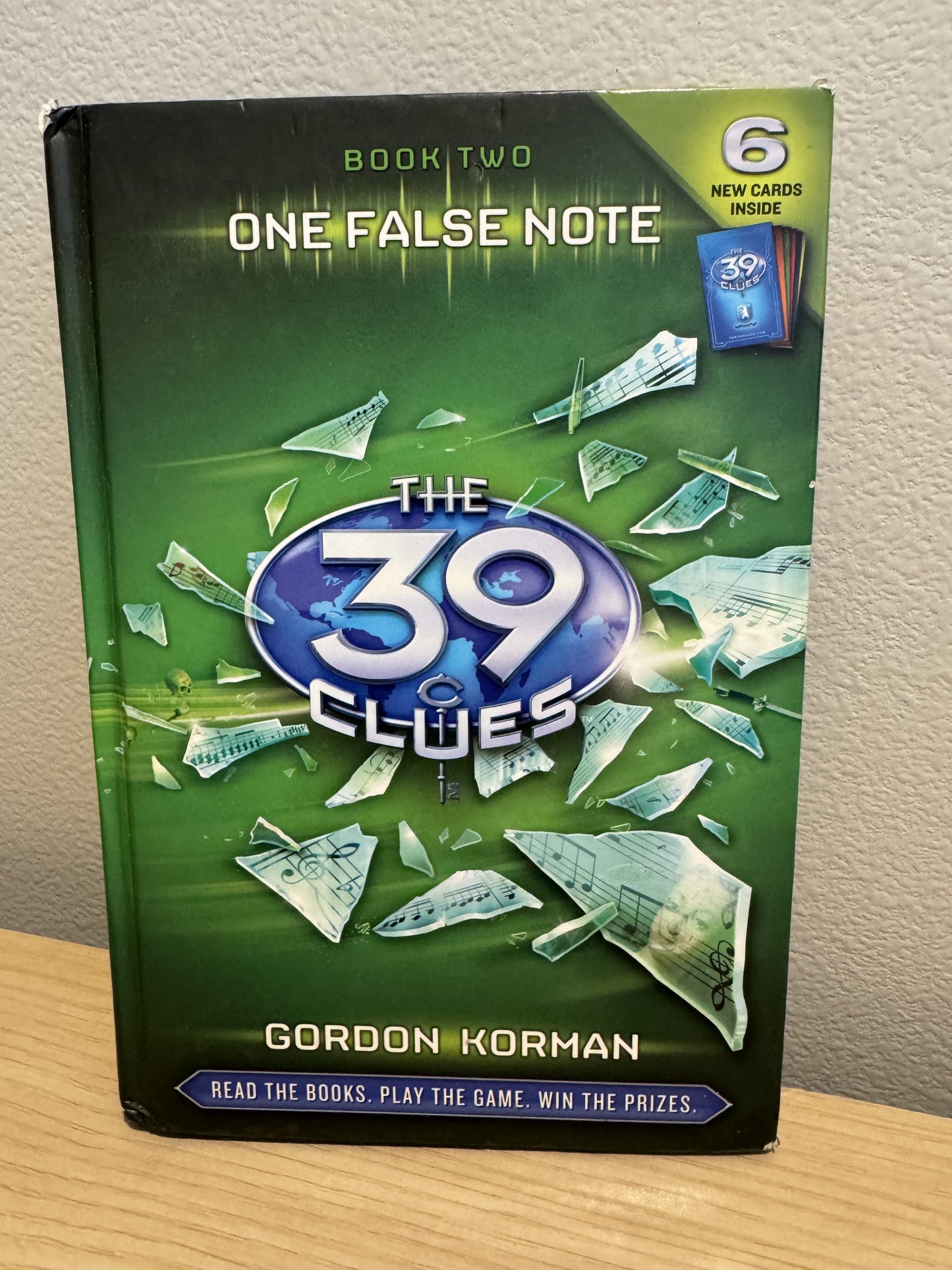 The 39 Clues: Book Two paperback game book by Gordon Korman, featuring a green cover with a blue and white title logo and scattered cash and note graphics, standing on a wooden surface against a beige wall.