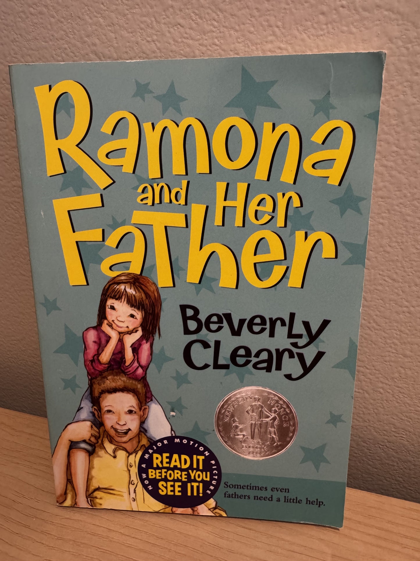 The book cover of 'Ramona and Her Father' by Beverly Cleary, featuring a teal background with yellow title text, an illustration of a girl sitting on a boy's shoulders, and a gold Medallion of the Reader award seal. The book is photographed standing on a wooden surface against a beige wall.