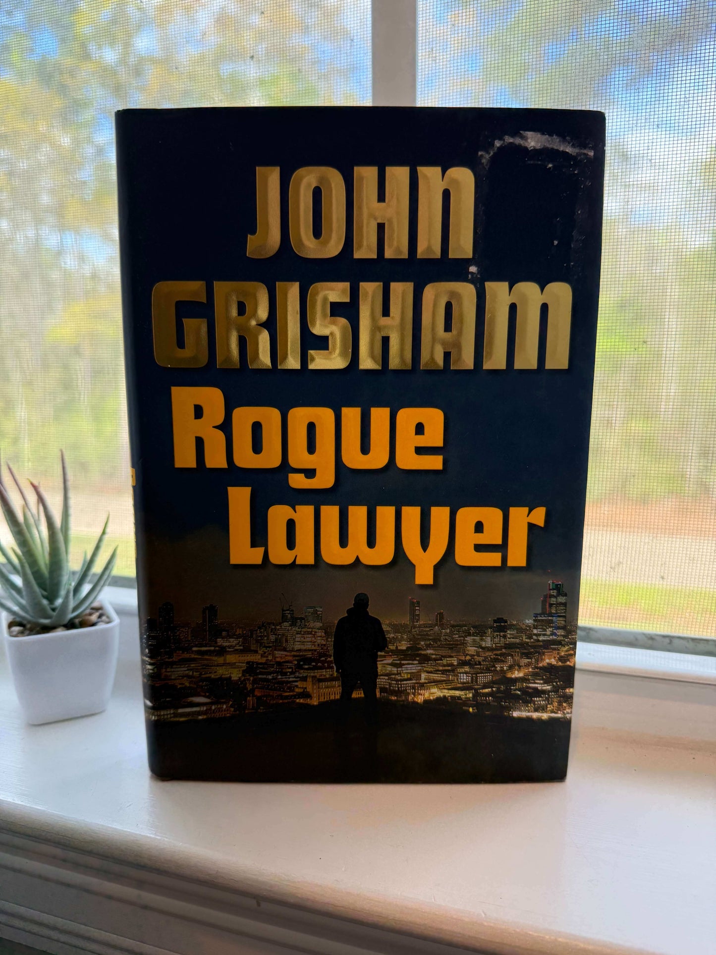Book titled 'Rogue Lawyer' by John Grisham on a windowsill with a view of trees outside.