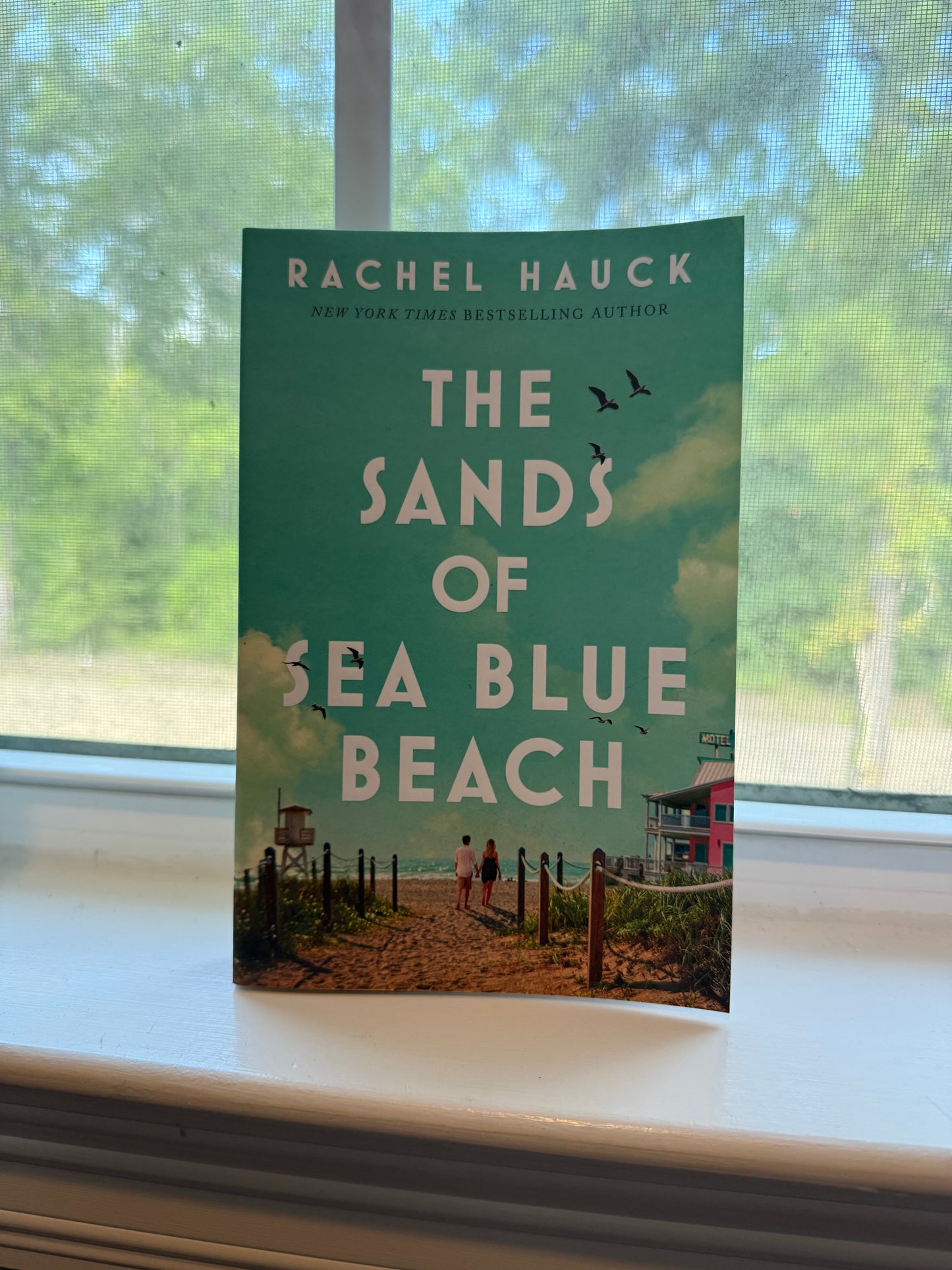 Book titled 'The Sands of Sea Blue Beach' by Rachel Hauck on a window sill with a blurred outdoor background.