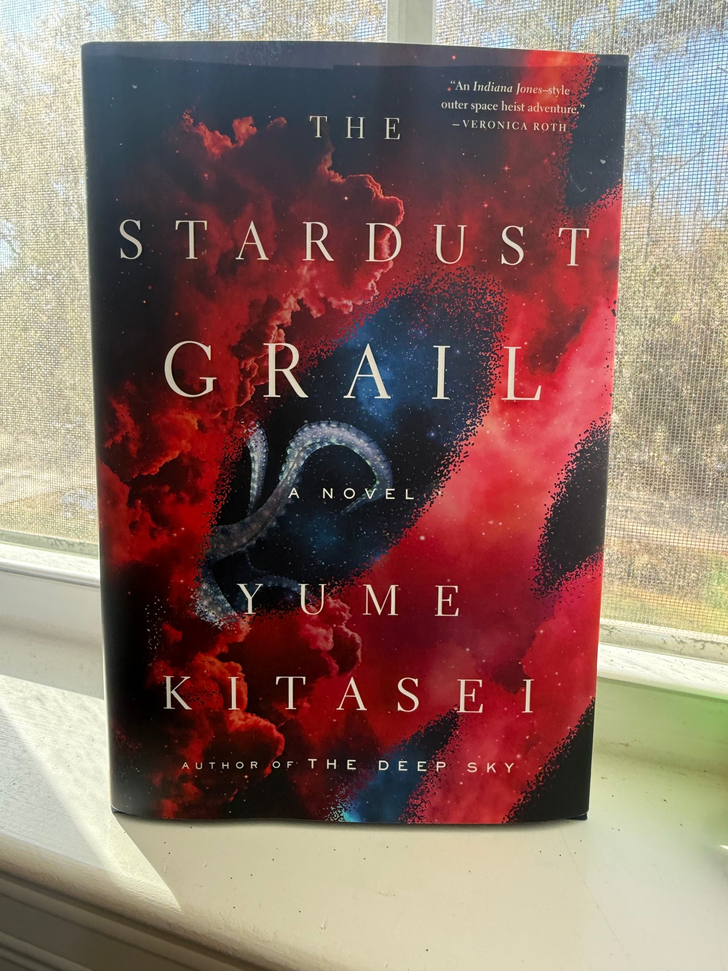 A book titled 'The Stardust Grail' by Yume Kitasei, featuring a vibrant cover with a blend of red and purple hues, placed on a windowsill.