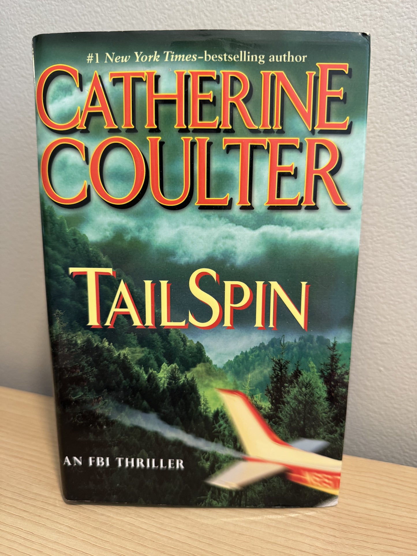 A paperback copy of the book 'Tailspin' by Catherine Coulter, featuring a dark green cover with red and yellow title text and an airplane scene near the bottom, photographed standing on a wooden surface against a light wall.