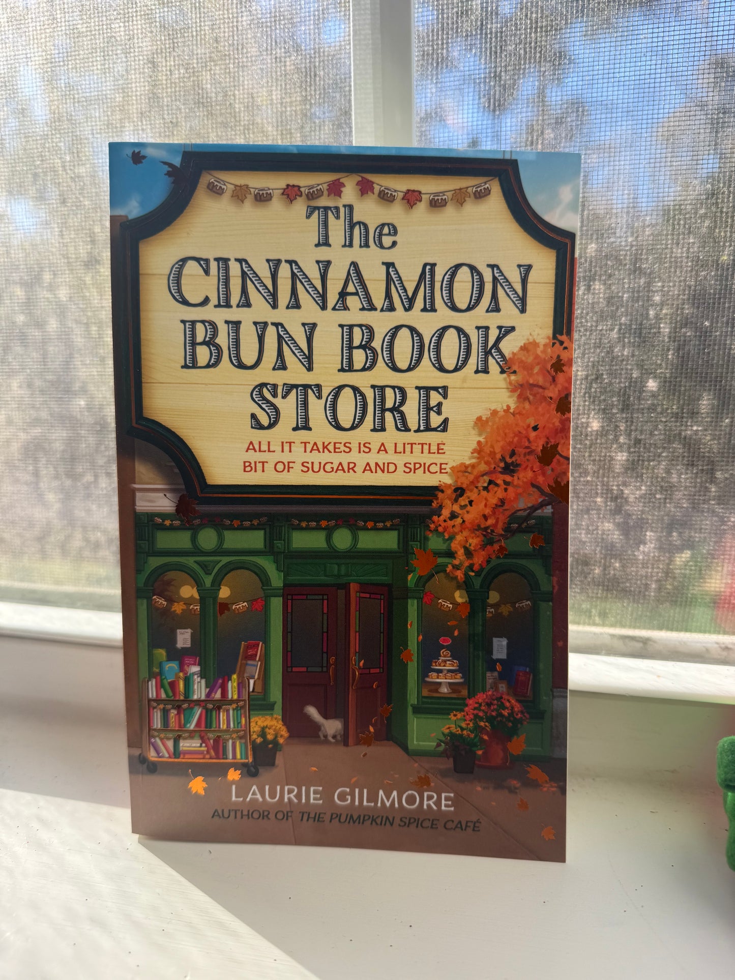 A book cover of 'The Cinnamon Bun Bookstore' by Lauren Gilmore, featuring an illustration of a bookstore with cinnamon buns, a cat, and autumn decorations.
