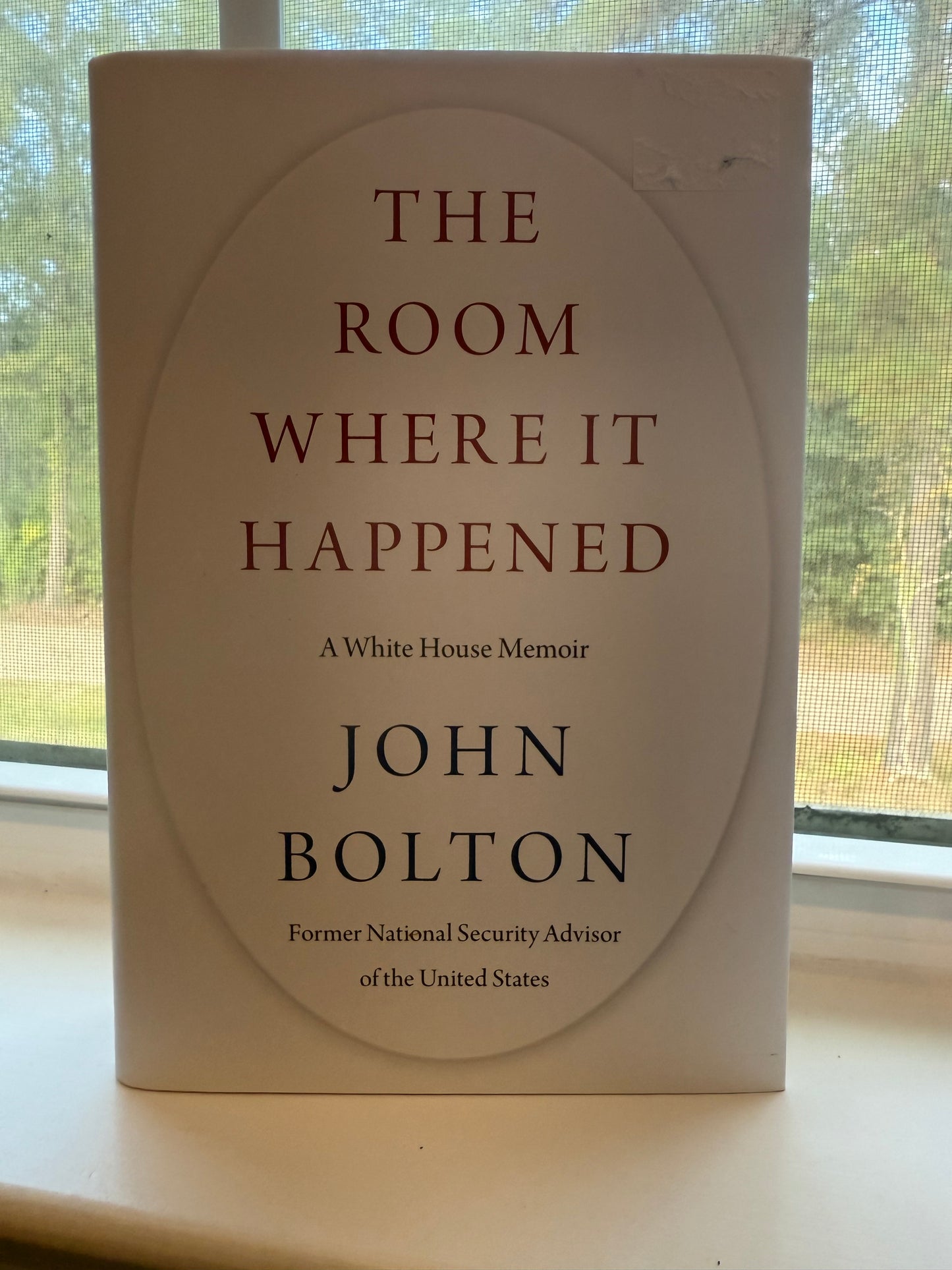 Book titled 'The Room Where It Happened' by John Bolton on a surface with a window in the background.