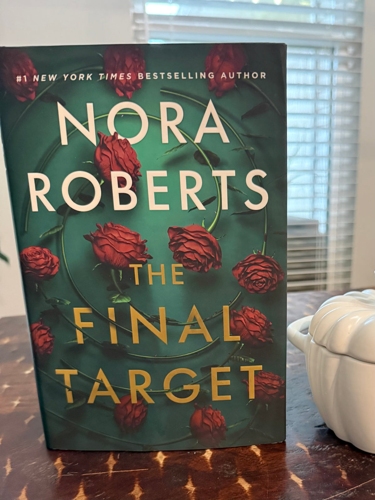 The book cover of Nora Roberts' 'The Final Target' is shown standing upright on a wooden table, with a white ceramic container and window blinds in the background.