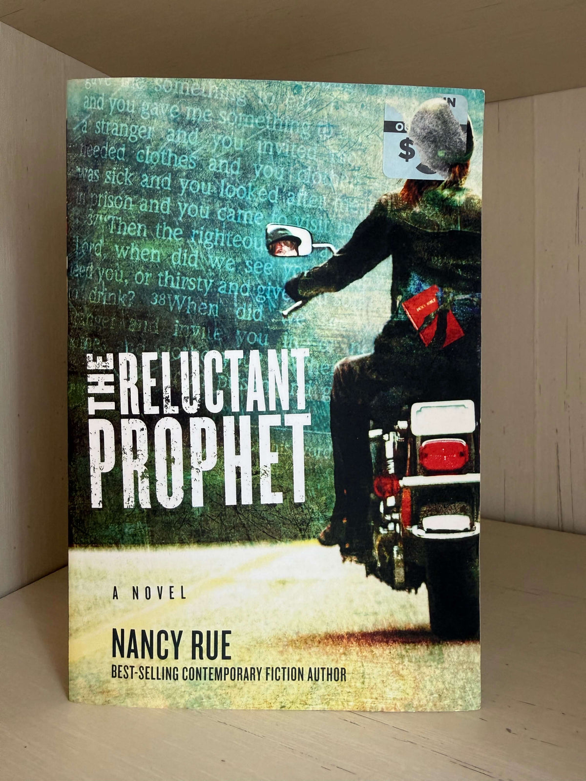 The book cover of 'The Reluctant Prophet' by Nancy Rue, featuring a stylized illustration of a person on a motorcycle with a dollar sign for a helmet, set against a textured teal background with text overlays.