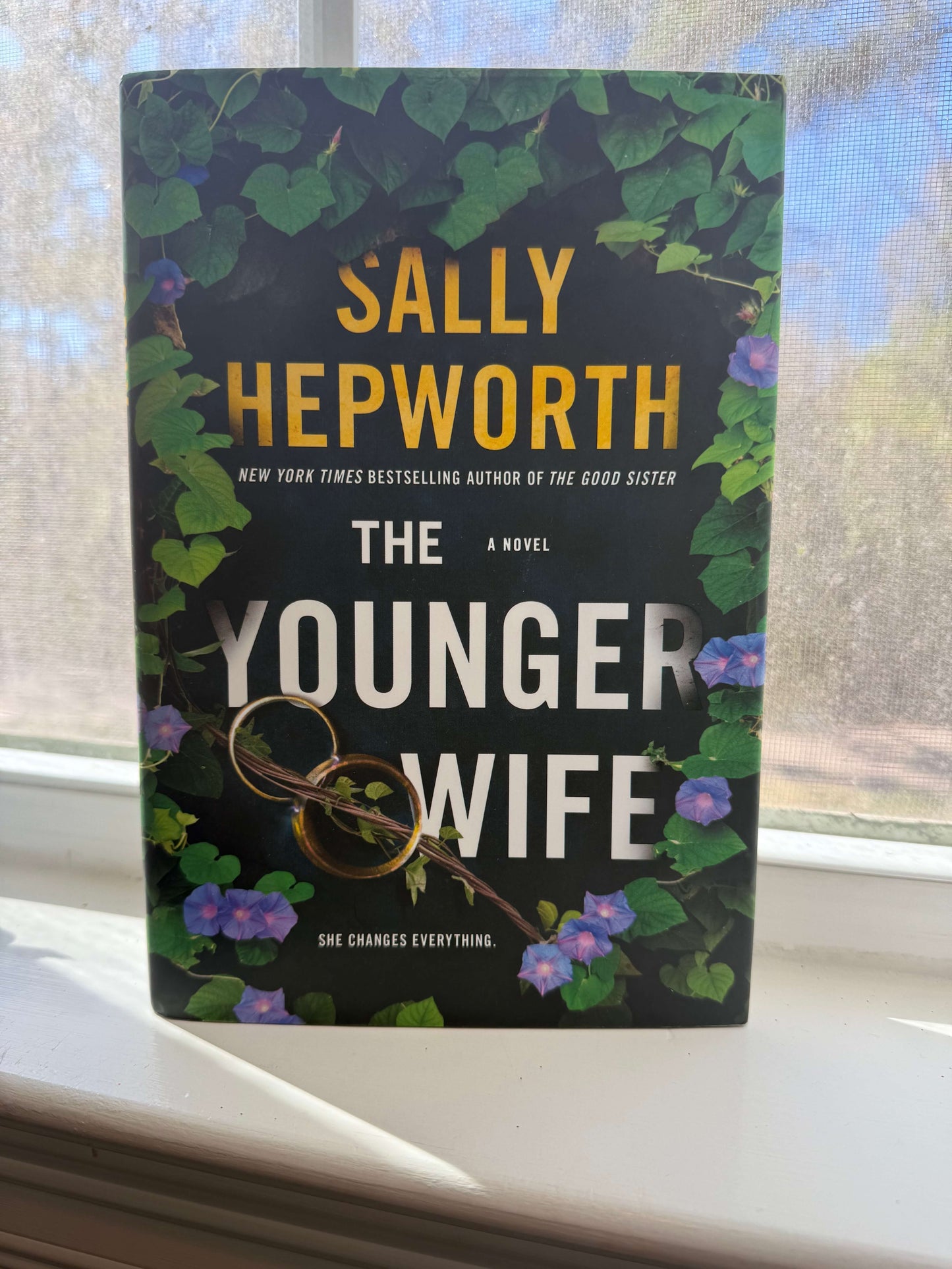 Book titled 'The Younger Wife' by Sally Hepworth on a windowsill with a blurred outdoor background.