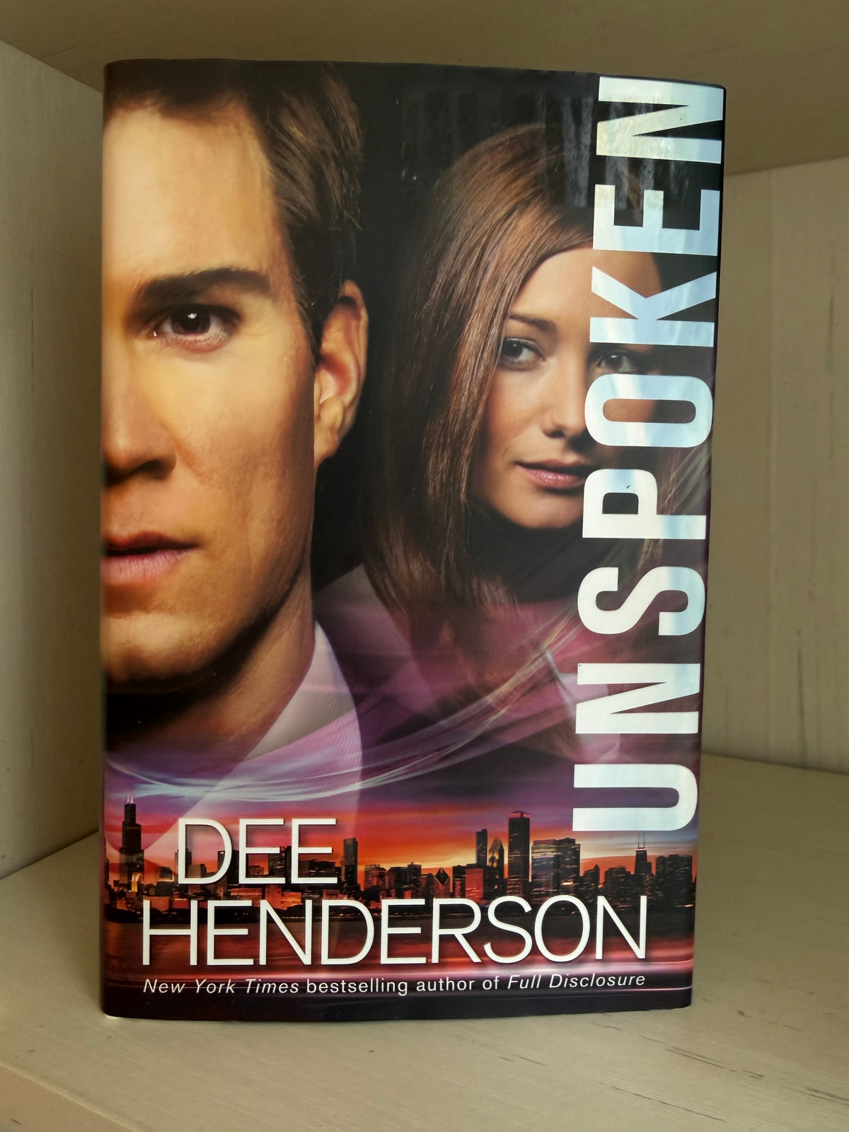 The book cover of 'Unspoken' by Dee Henderson, featuring a close-up portrait of a man on the left, a woman on the right, and a city skyline at sunset along the bottom, photographed standing upright on a shelf.