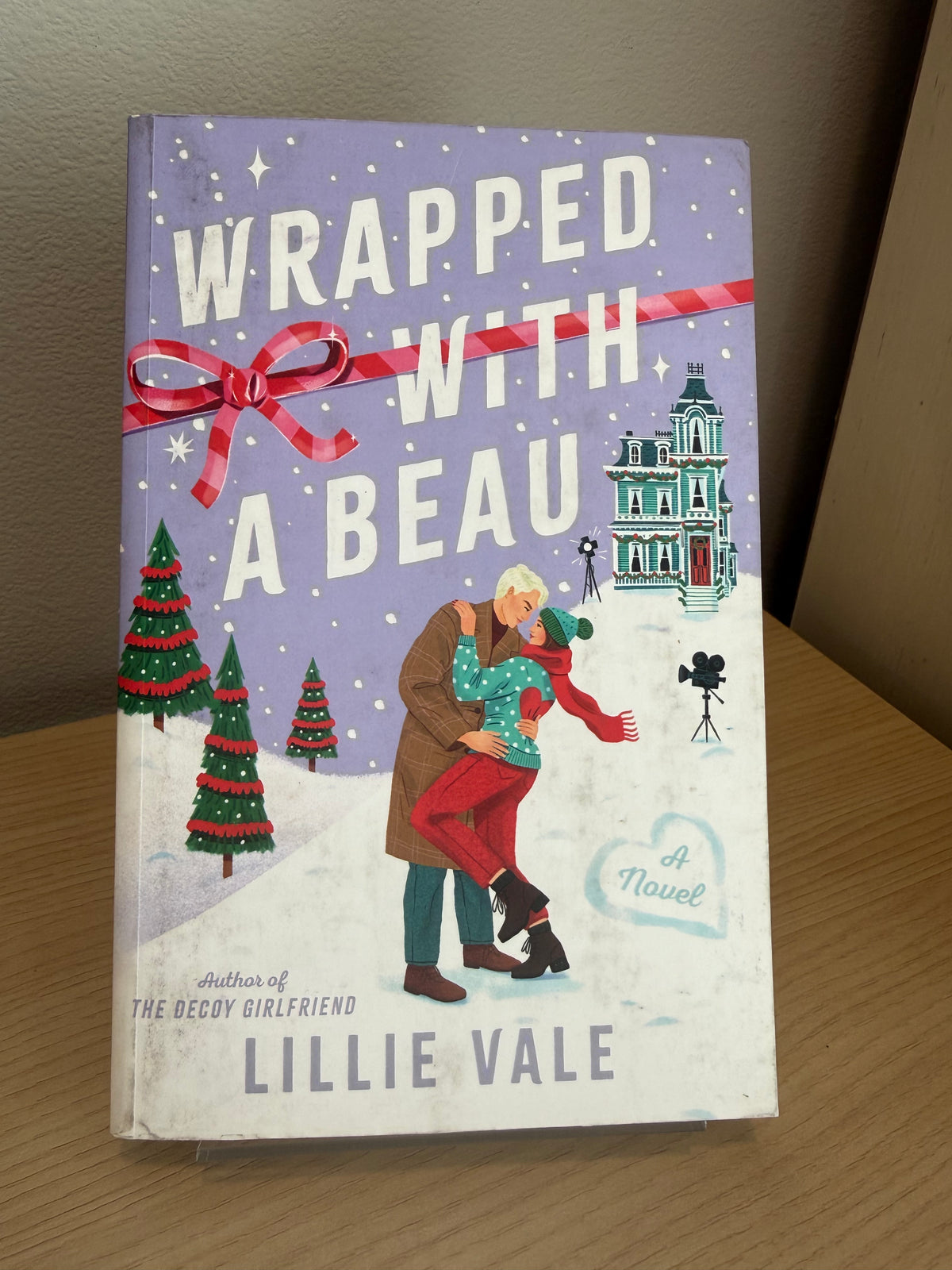 The book cover of 'Wrapped with a Beauty' by Lillie Vale, featuring a festive winter illustration with a couple embracing, Christmas trees, a house, and a red ribbon bow on a purple background, displayed on a wooden surface.