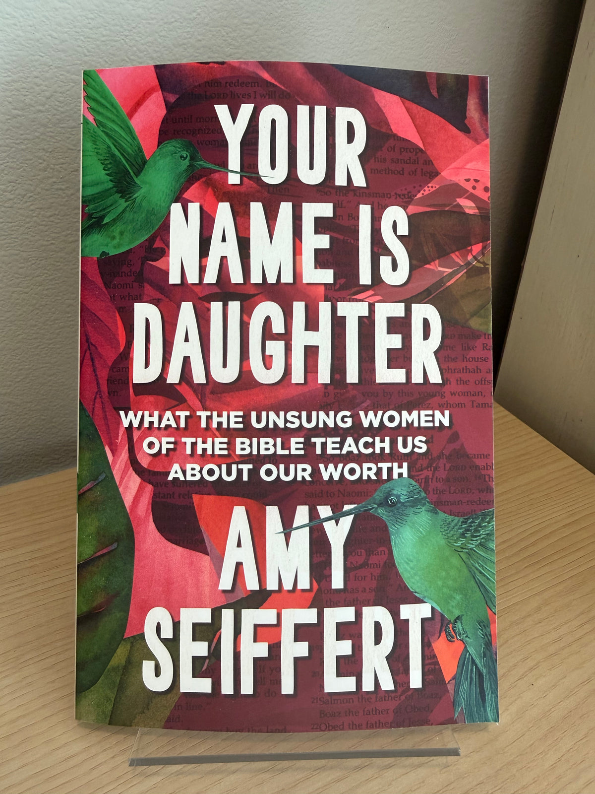 The book cover of 'Your Name Is Daughter' by Amy Seiffert, featuring a vibrant red and pink background with green hummingbirds and bold white text, displayed on a wooden surface.