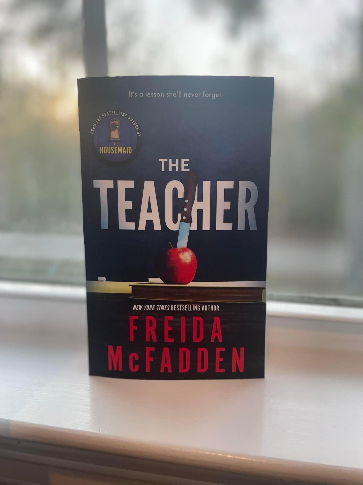The cover of the book 'The Teacher' by Freida McFadden features a stylized image of a hand holding a knife, with a apple and a book, against a dark background.