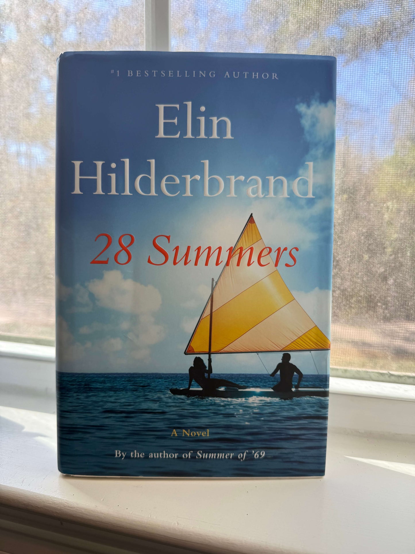 Book titled '28 Summers' by Elin Hilderbrand on a windowsill with a view of water and sky.
