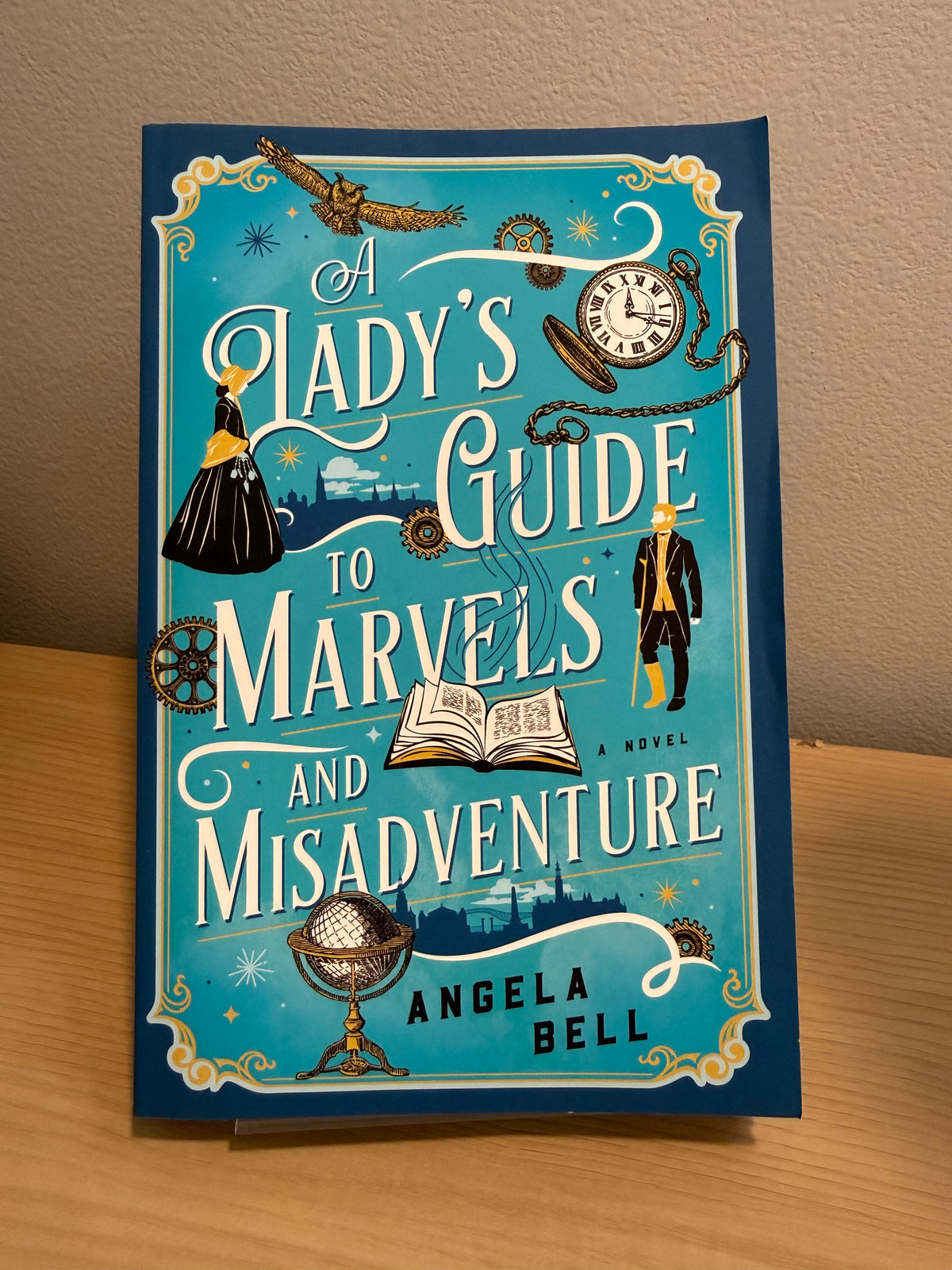A book titled 'A Lady's Guide to Marvels and Misadventure' by Angela Bell, standing upright on a wooden surface against a beige wall. The cover features a teal background with vintage illustrations of a clock, globe, book, and figures, along with ornate gold decorative borders.
