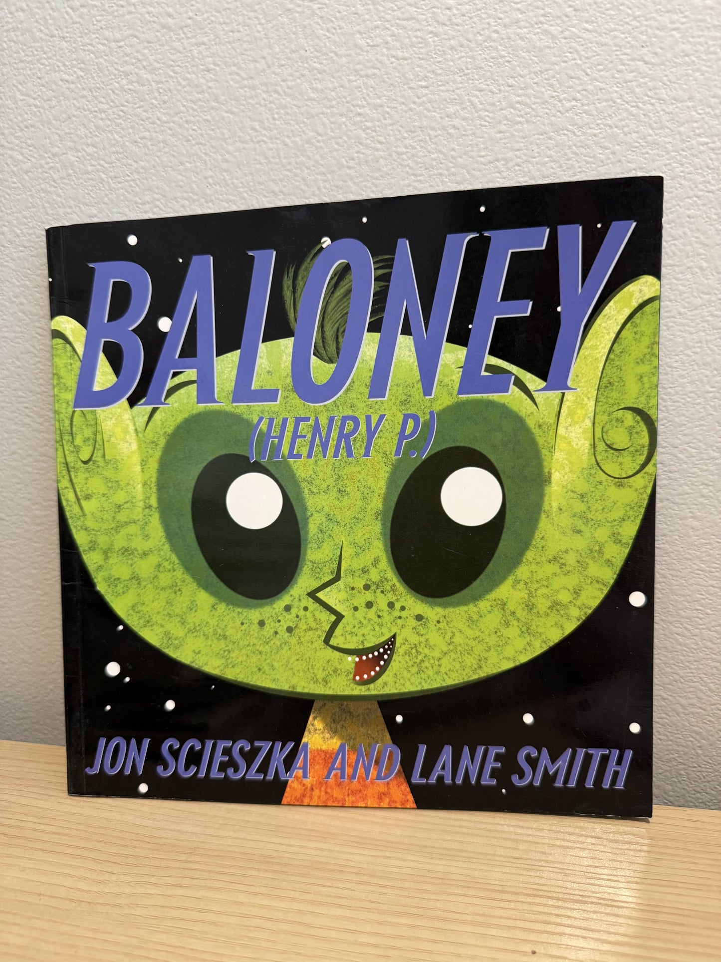 The front cover of the children's book 'Baloney (Henry P.)' by Jon Scieszka and Lane Smith, featuring a green alien face on a black starry background, leaning against a light gray wall on a light wood surface.