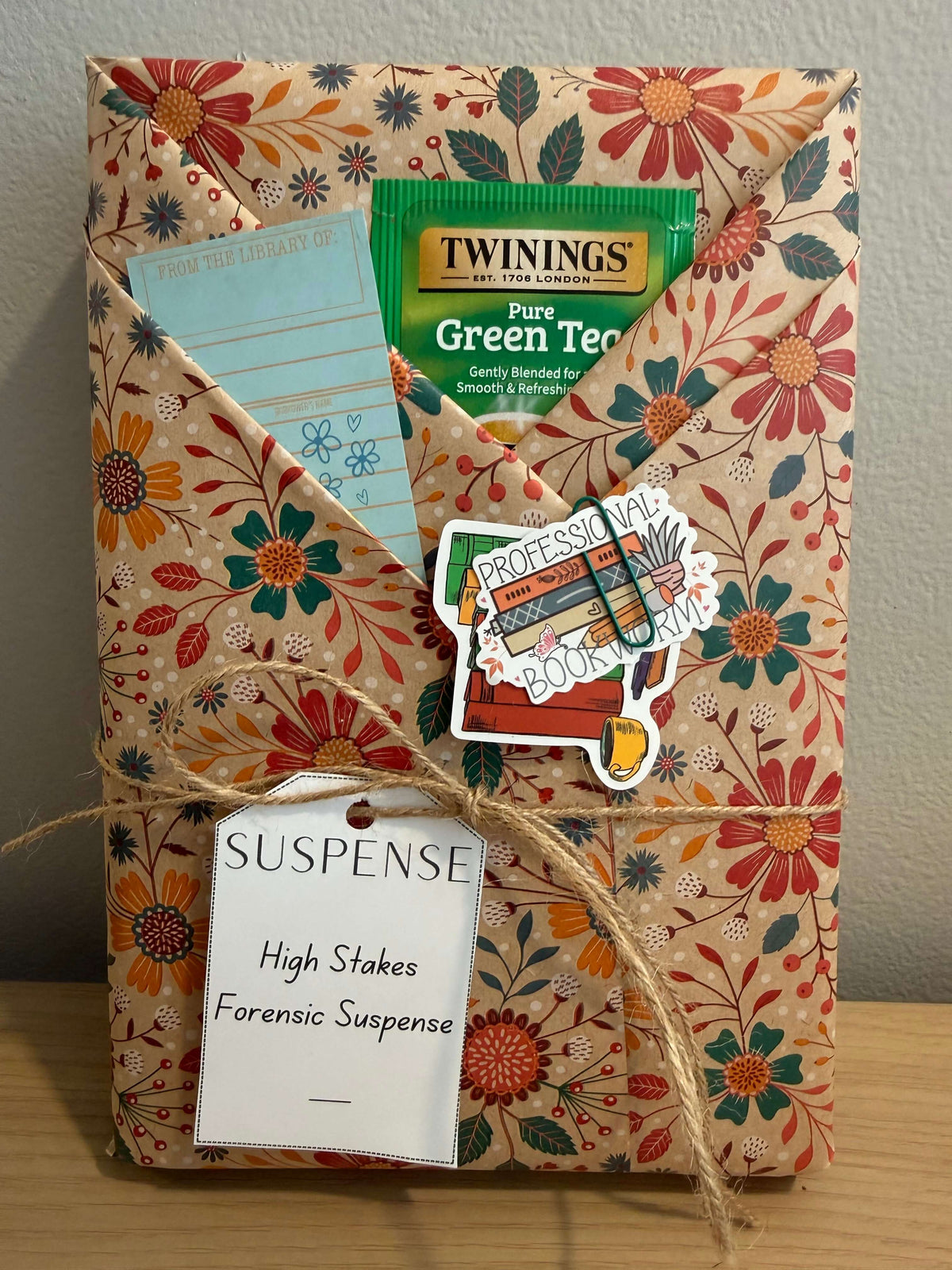 A wrapped gift box with a floral envelope-style wrap in beige, orange, red, and teal tones, tied with twine and a paper tag reading 'SUSPENSE High Stakes Forensic Suspense.' A Twinings Pure Green Tea packet is tucked into the top flap, along with a blue card and a decorative sticker that says 'Professors of Bookworms.