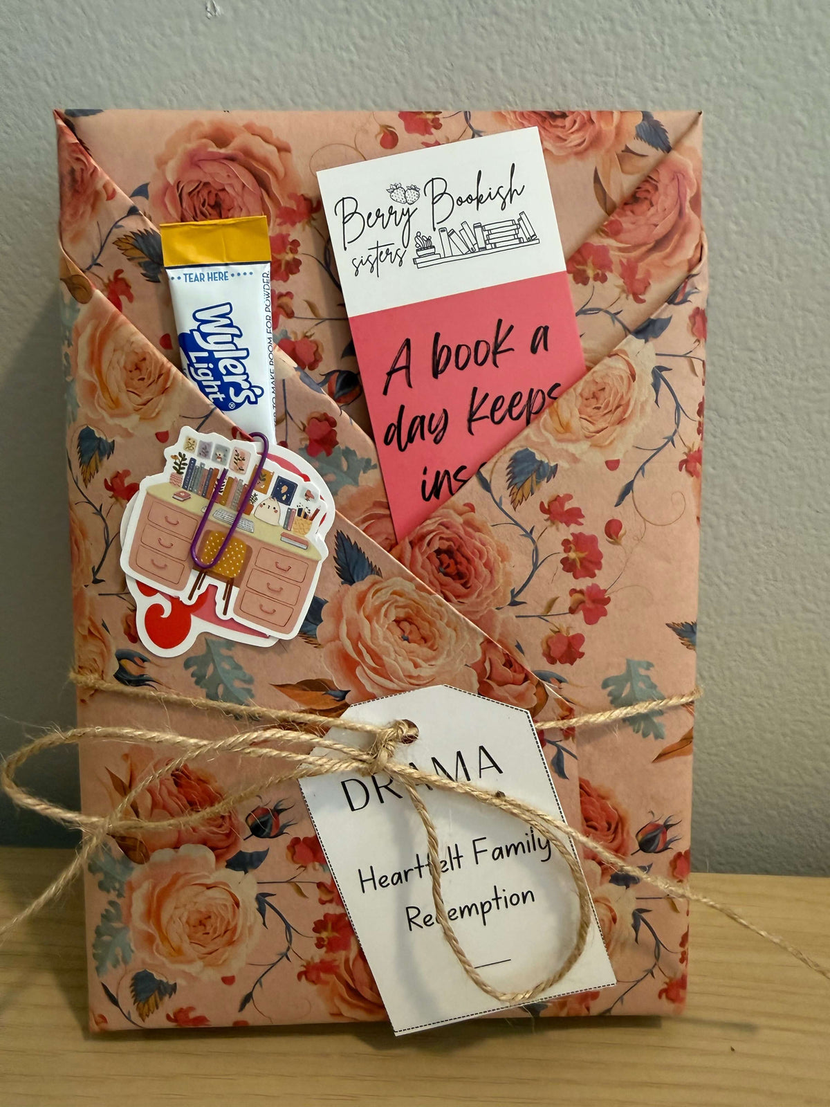 A handmade gift package wrapped in peach floral paper with a pink rose pattern, tied with twine and decorated with stickers, a small Wilton's Lighters tube, and handwritten tags.