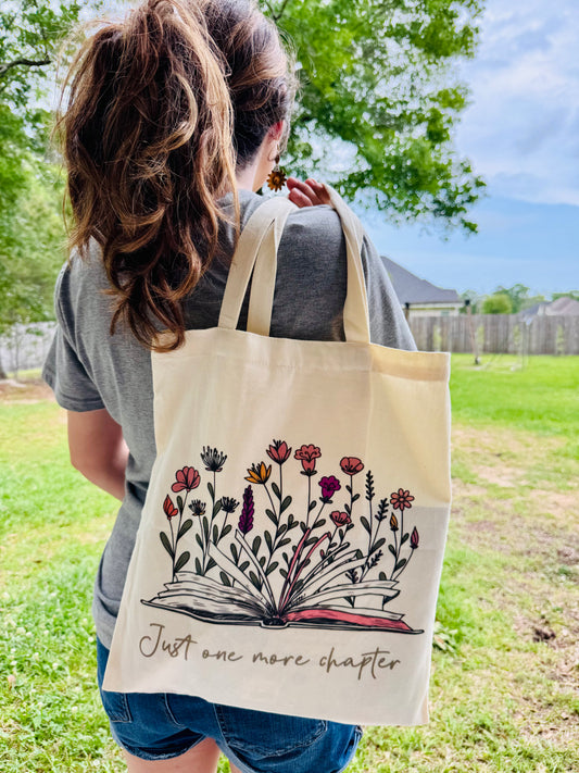 Person holding a tote bag with floral design and text 'Just one more chapter' outdoors.