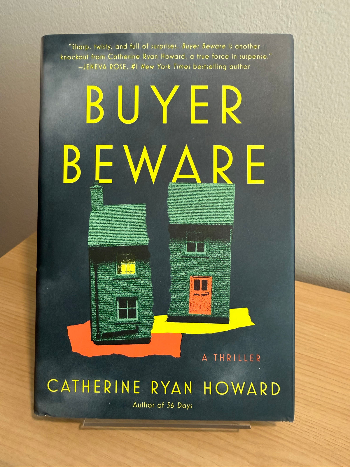 The book 'Buyer Beware' by Catherine Ryan Howard, featuring a dark gray cover with bold yellow title text and an illustration of two green houses, displayed on a wooden surface.
