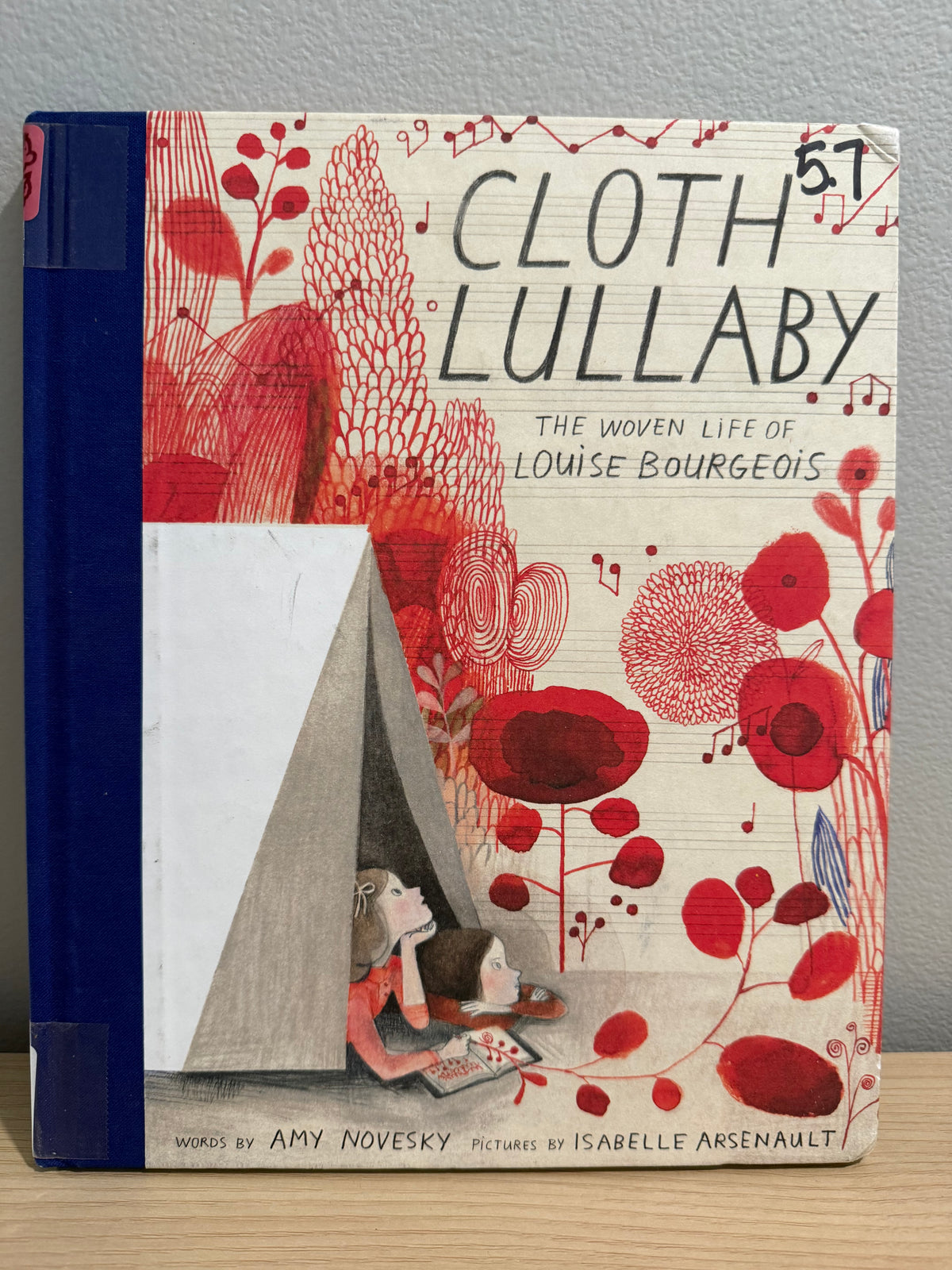 The book 'Cloth Lullaby' by Amy Novesky, featuring a cream-colored cover with red floral illustrations and a navy blue spine, displayed on a wooden surface against a gray wall.