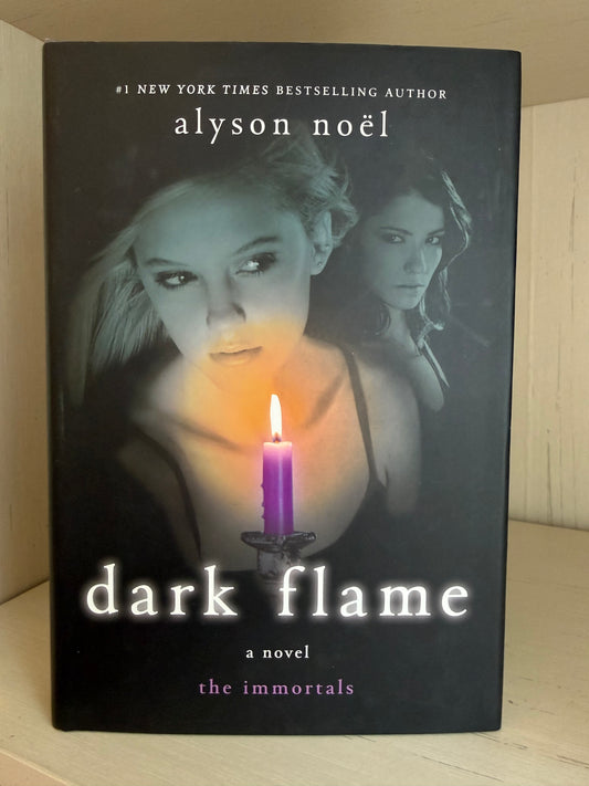 The book cover of 'Dark Flame: A Novel' by Alyson Noel, featuring a dark background with a glowing purple candle and two women's faces, photographed standing upright on a light wood surface.