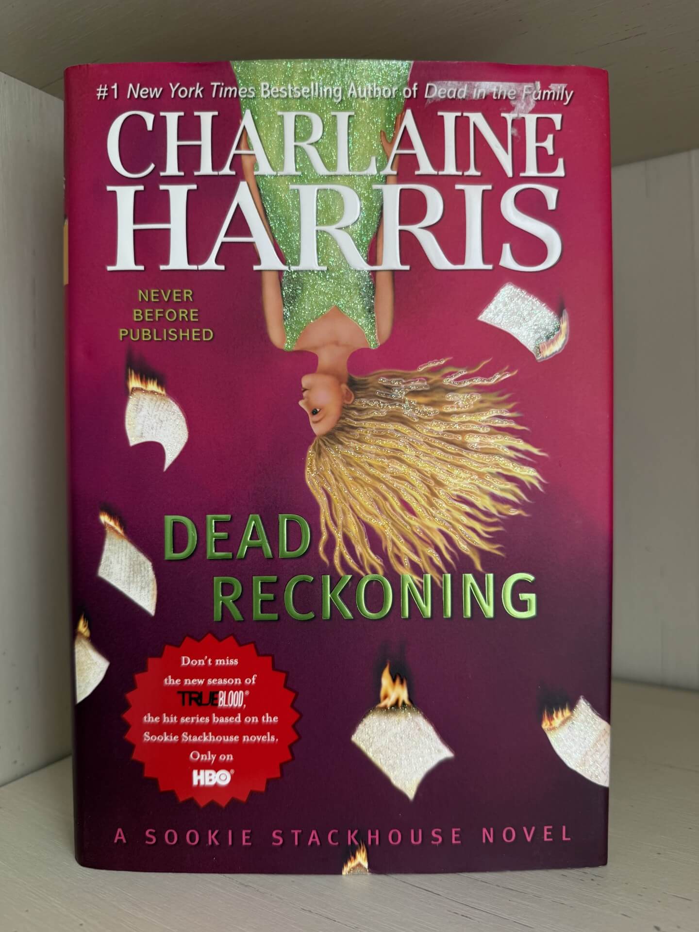 A hardcover book titled 'Dead Reckoning' by Charlaine Harris, photographed standing upright against a light gray wall. The cover is predominantly deep magenta/purple with large white author text, green title text, and an illustration of a woman with flowing blonde hair surrounded by floating papers and small flames.