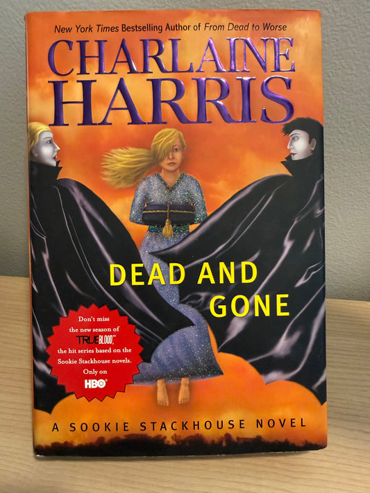 A hardcover book of 'Dead and Gone' by Charlaine Harris, featuring a vibrant orange and black cover illustration with a central figure in a blue dress, placed on a wooden surface against a gray wall.