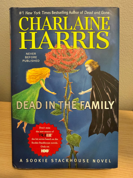 A hardcover book of Charlaine Harris's 'Dead in the Family' novel, featuring a dark blue cover with a large red rose, a blonde woman in a green dress, and a woman in a black cloak. The author's name is printed in large yellow letters at the top.