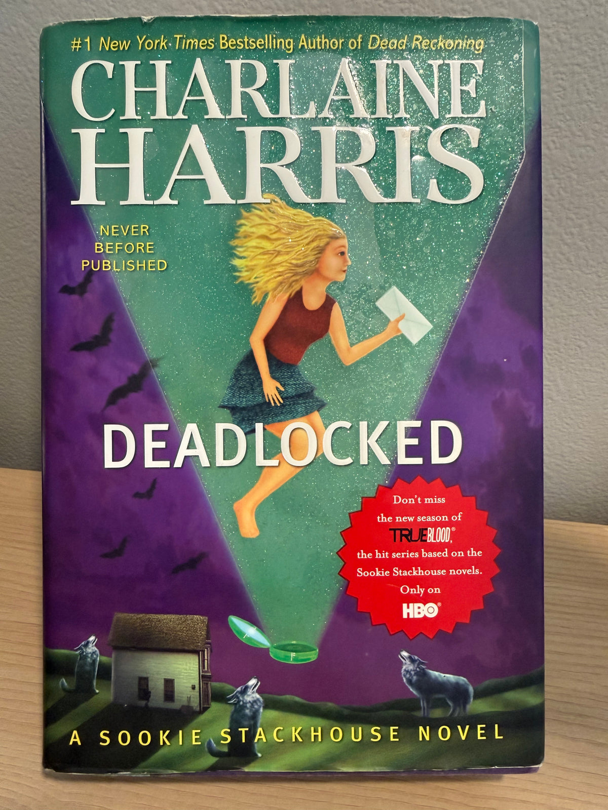 Book cover of the novel 'Deadlocked' by Charlaine Harris, featuring a purple and teal background with an illustration of a woman running, wolves, a small house, and a red circular promotional badge.