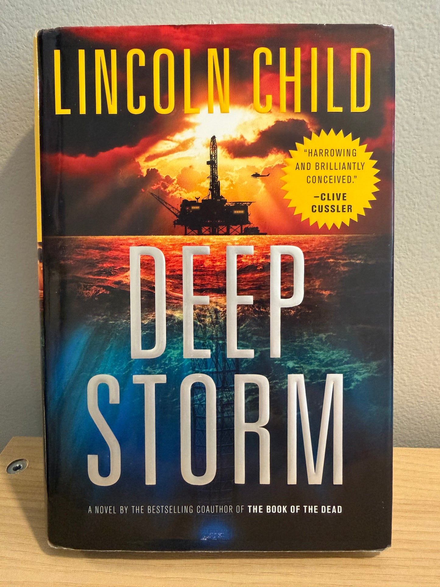 A hardcover book titled "Deep Storm" by Lincoln Child, photographed from above on a light wood surface against a light wall. The cover features a dramatic storm scene with red, orange, blue, and black tones, large white title text, and a yellow burst quote near the top left.