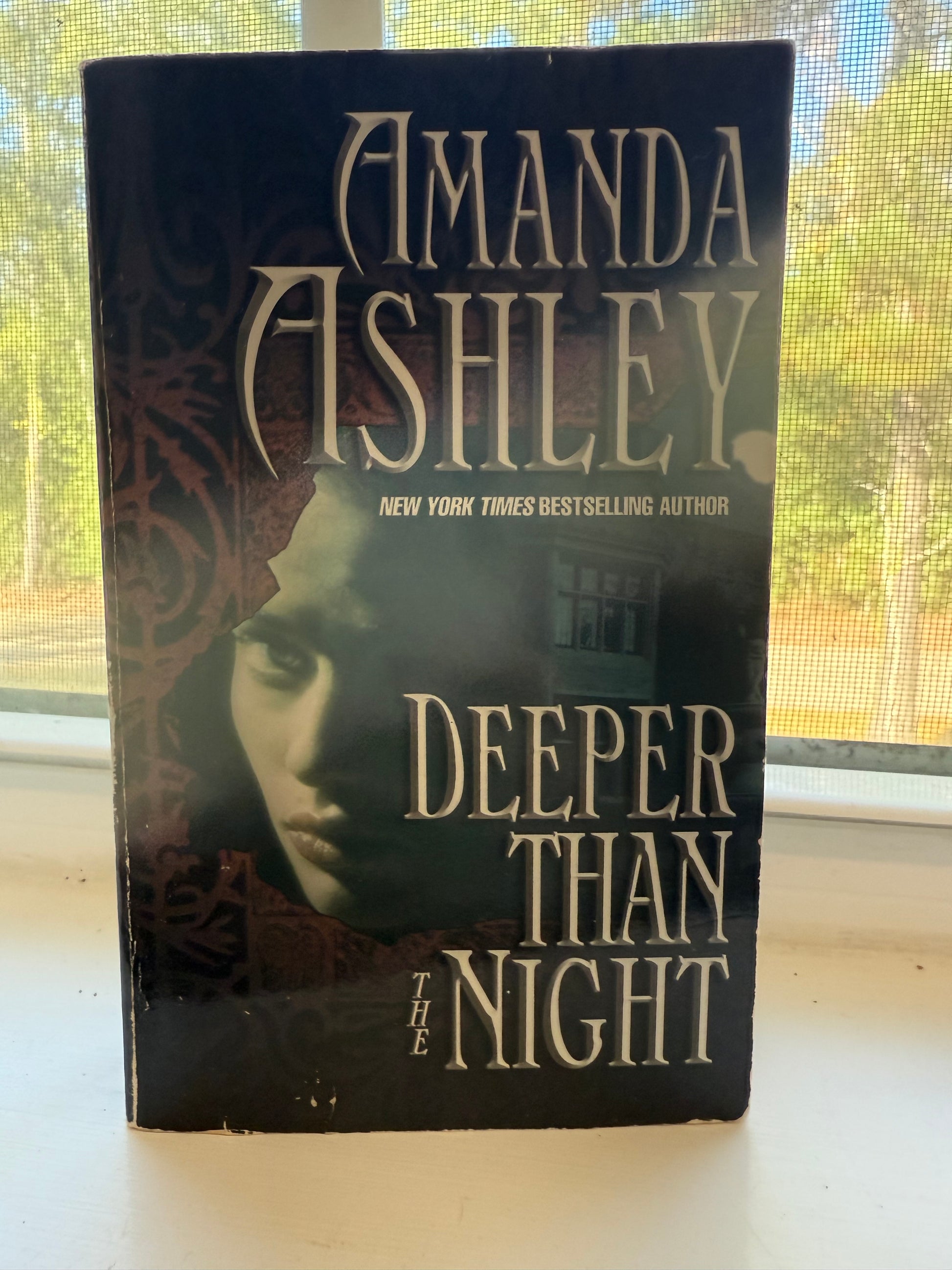 Book titled 'Deeper Than Night' by Amanda Ashley on a surface with a window in the background.