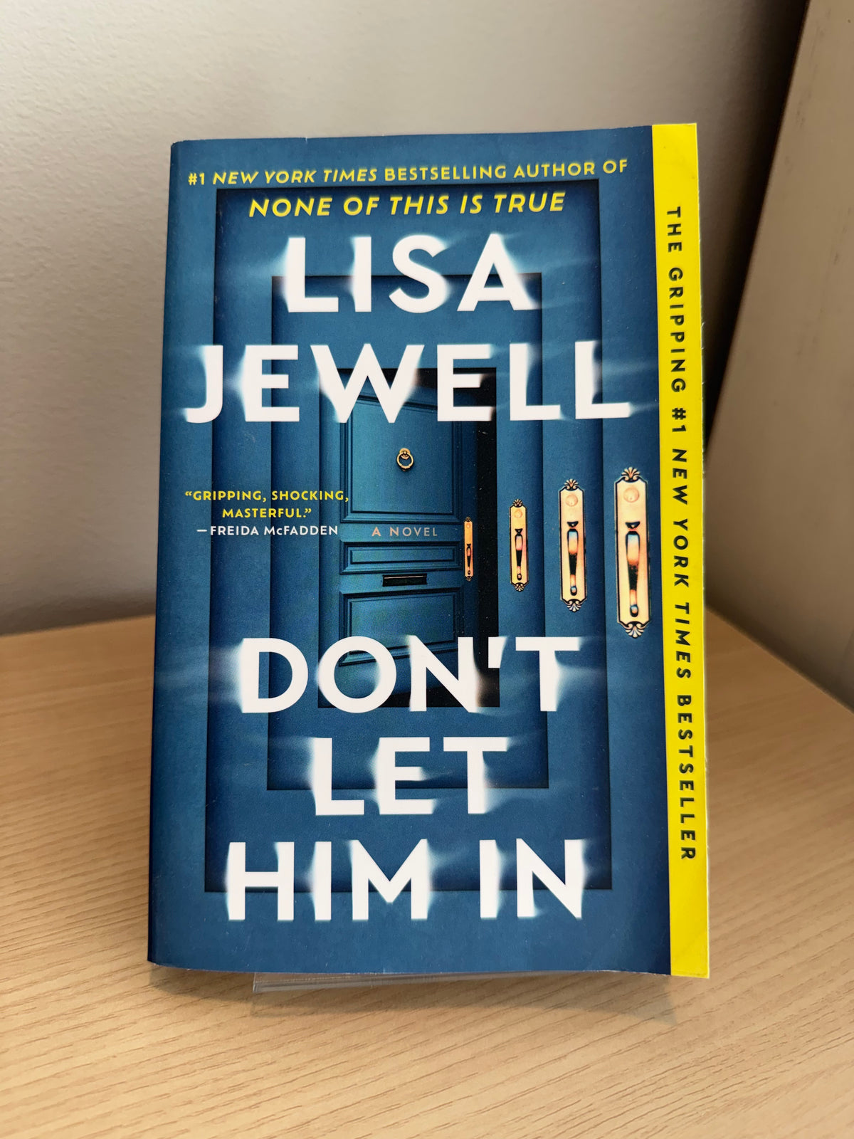 The gripping, shocking bestseller novel 'Don't Let Him In' by Lisa Jewell, shown standing upright on a small clear display block on a light wood surface.