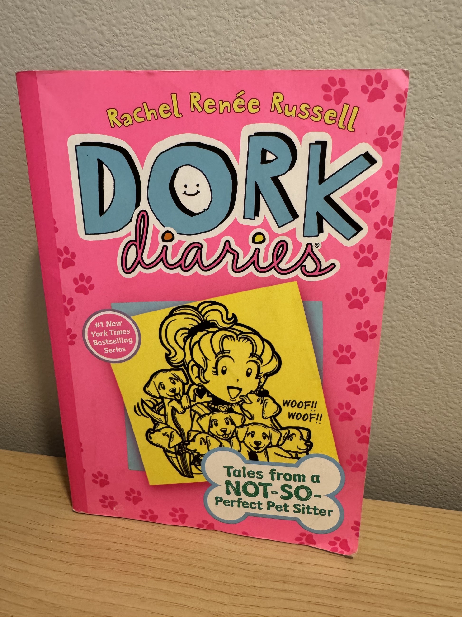 The book 'Dork Diaries: Tales from a Not-So-Perfect Pet Sitter' by Rachel Renée Russell, featuring a bright pink cover with cartoon dog paw prints and a yellow illustration of a girl with dogs, shown standing on a wooden surface against a textured wall.