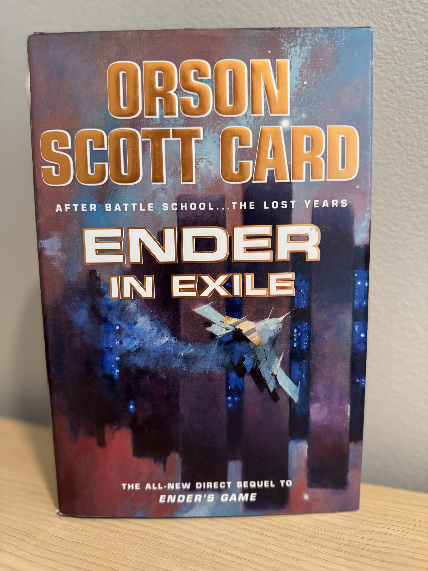 The book cover of 'Ender in Exile' by Orson Scott Card, standing upright on a light wood surface against a neutral wall.