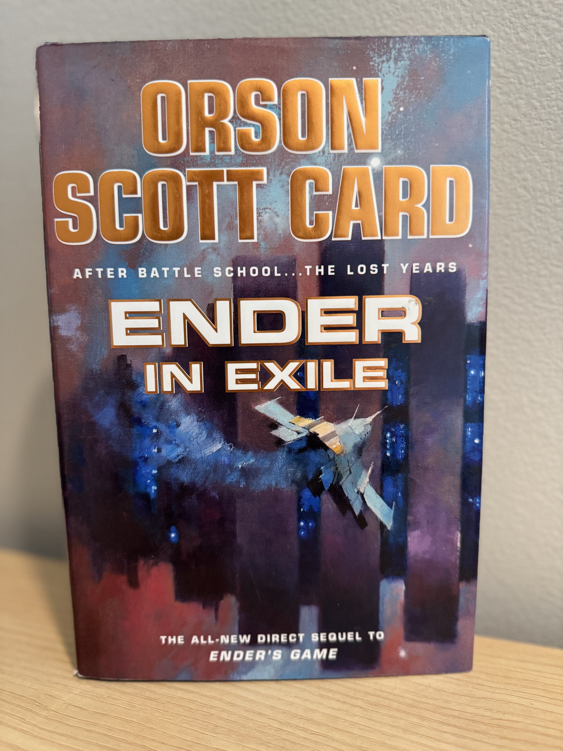 The book cover of 'Ender in Exile' by Orson Scott Card, standing upright on a light wood surface against a neutral wall.