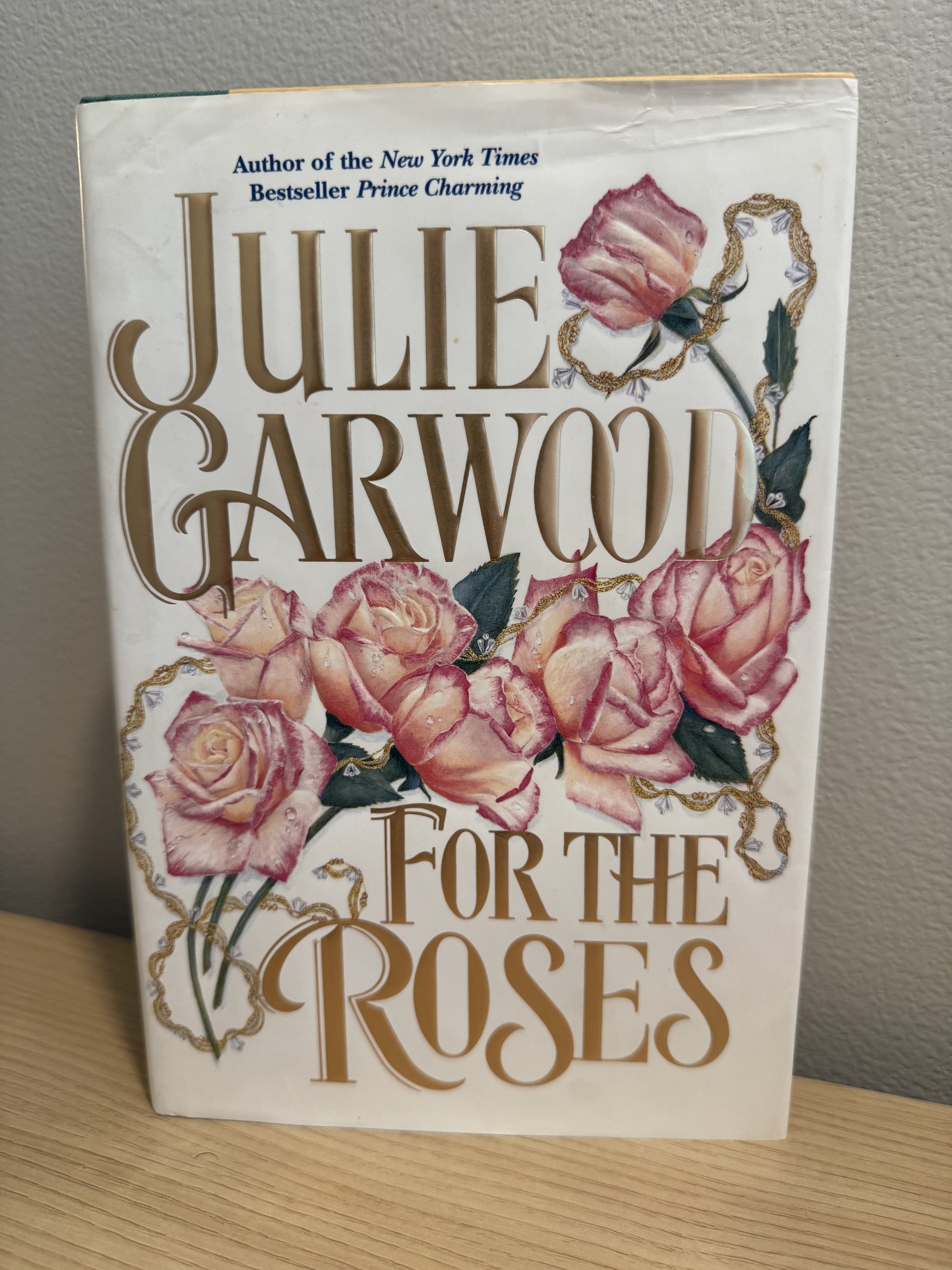 The front cover of the book 'For the Roses' by Julie Garwood, featuring pink roses and gold lettering on an off-white background, photographed standing on a wooden surface against a gray wall.