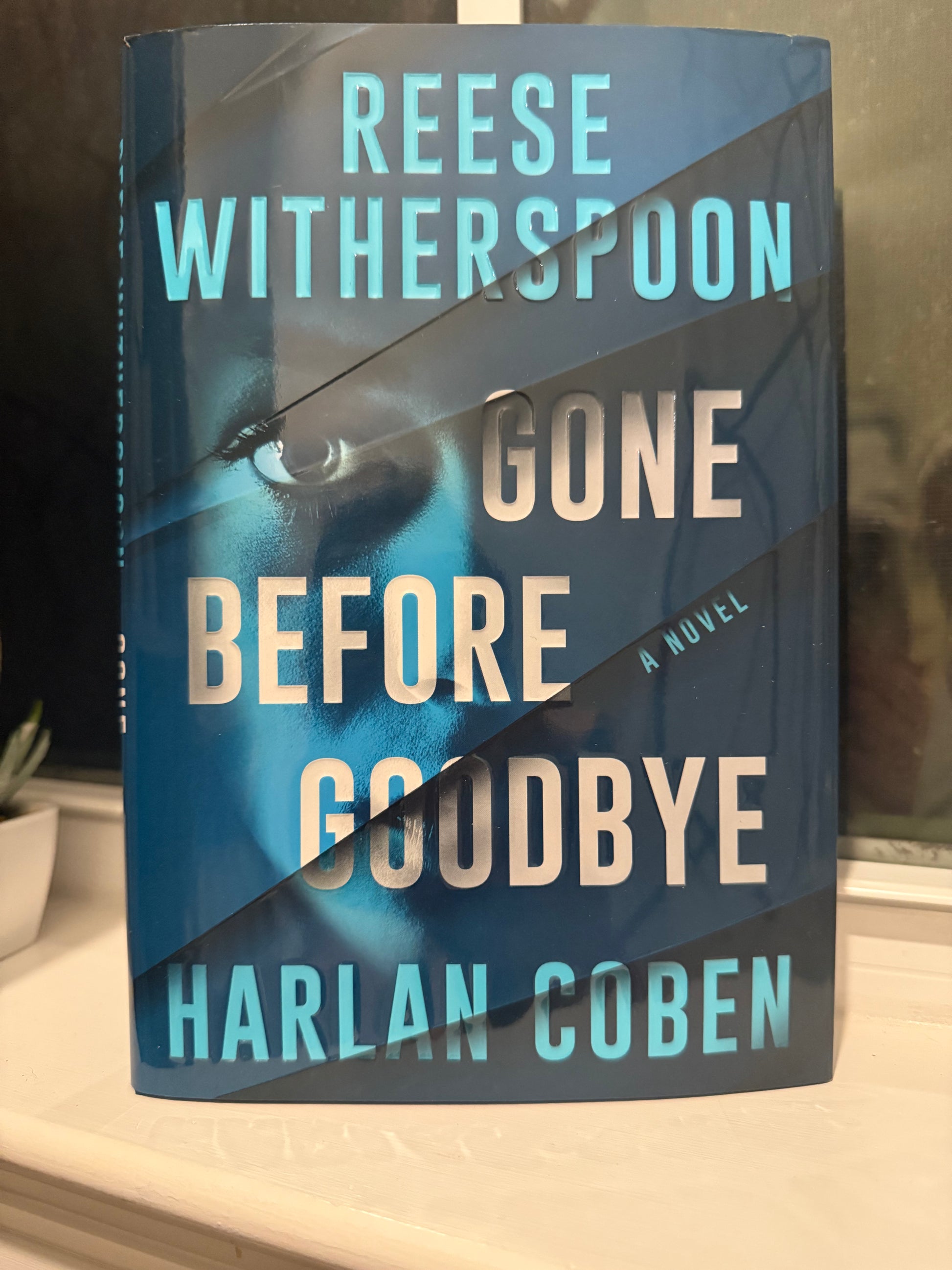 Book titled 'Before Goodbye' by Harlan Coben and 'Gone' by Reese Witherspoon on a reflective surface.