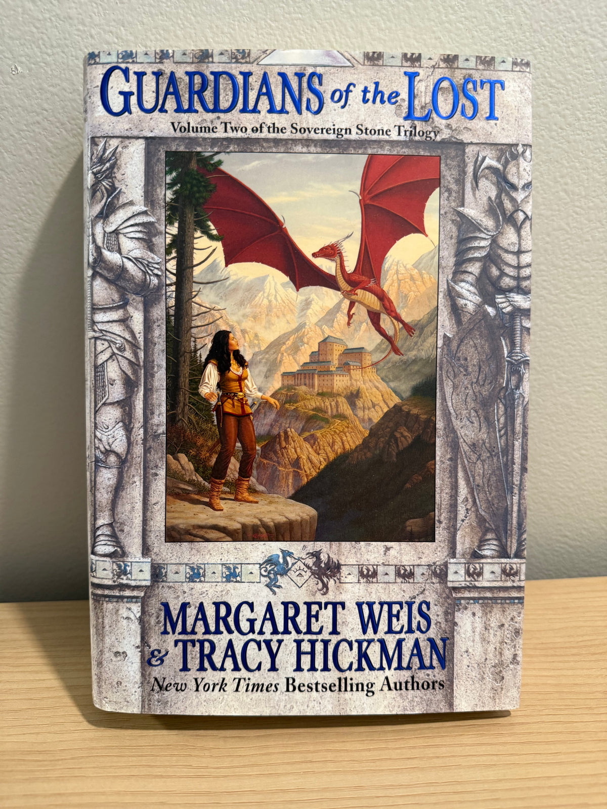 The book 'Guardians of the Lost' by Margaret Weis and Tracy Hickman, featuring a fantasy illustration of a warrior facing a red dragon on a stone wall, set on a light wood surface against a neutral wall.