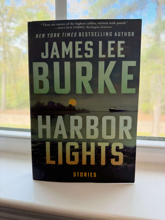 Book titled 'Harbor Lights' by James Lee Burke on a windowsill with a view of trees outside.