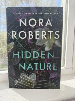 Book titled 'Hidden Nature' by Nora Roberts on a surface with a blurred window background
