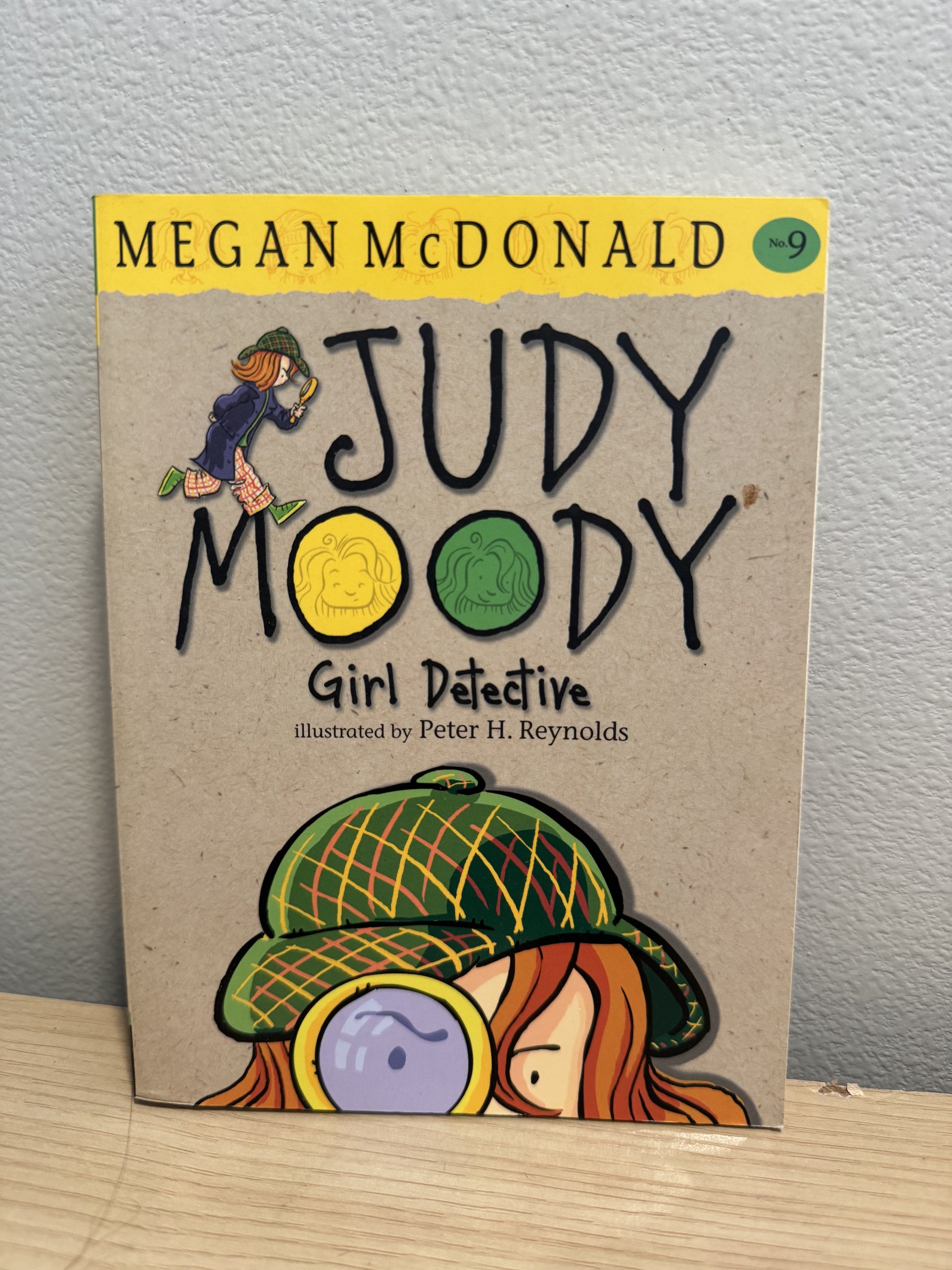 The front cover of the children's book 'Judy Moody Girl Detective' by Megan McDonald, featuring a beige background with a yellow banner at the top and cartoon illustrations of Judy Moody in a green plaid hat and magnifying glass.